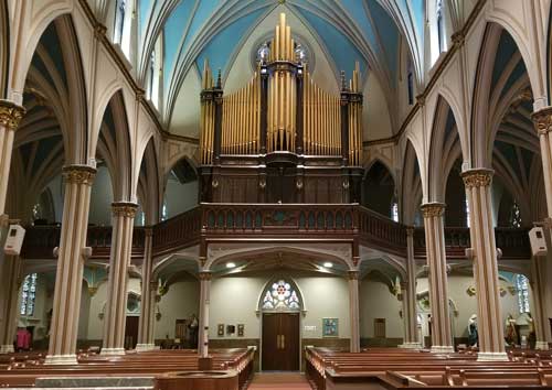 Interior view of a cathedral featuring a large pipe organ above a wooden gallery. The vaulted ceiling with blue accents and arched pillars create a grand and historic atmosphere. Pews line the sides leading towards the altar area.