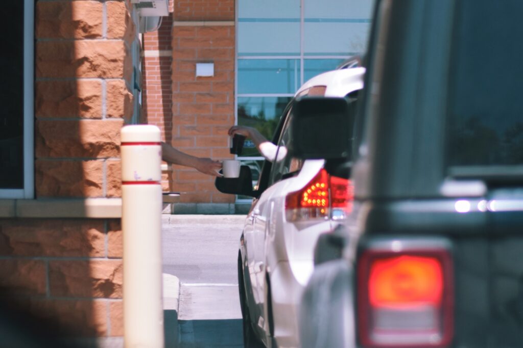Processed with VSCO with k3 preset A person hands a cup through the drive-thru window to someone in a white car, while another dark vehicle waits behind in line. The scene is outside a brick building on a sunny day. Drive-thru Trends