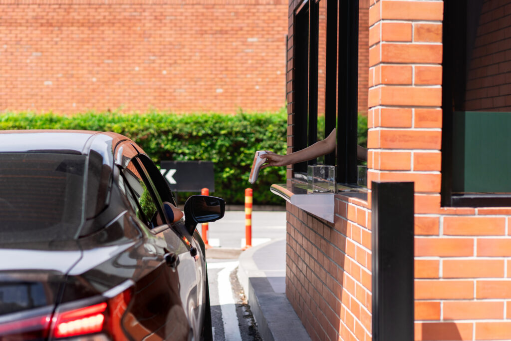 Fast and easy, scan to pay, pick up at the drive-thru, and enjoy your meal at home or at work. A person in a car receives food from a worker’s outstretched arm at a drive-thru window of a brick building. Green bushes and a brick wall are visible in the background. Drive-thru Trends.