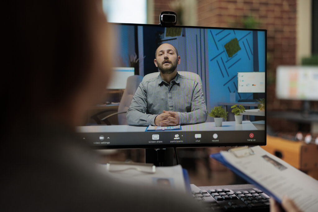 Businesswomen reading cv discussing application plan A person participates in a video call on a computer, with a man visible on the screen sitting at a desk. The foreground shows part of another person holding a clipboard and taking notes. Teams vs. Zoom. TSI Global.
