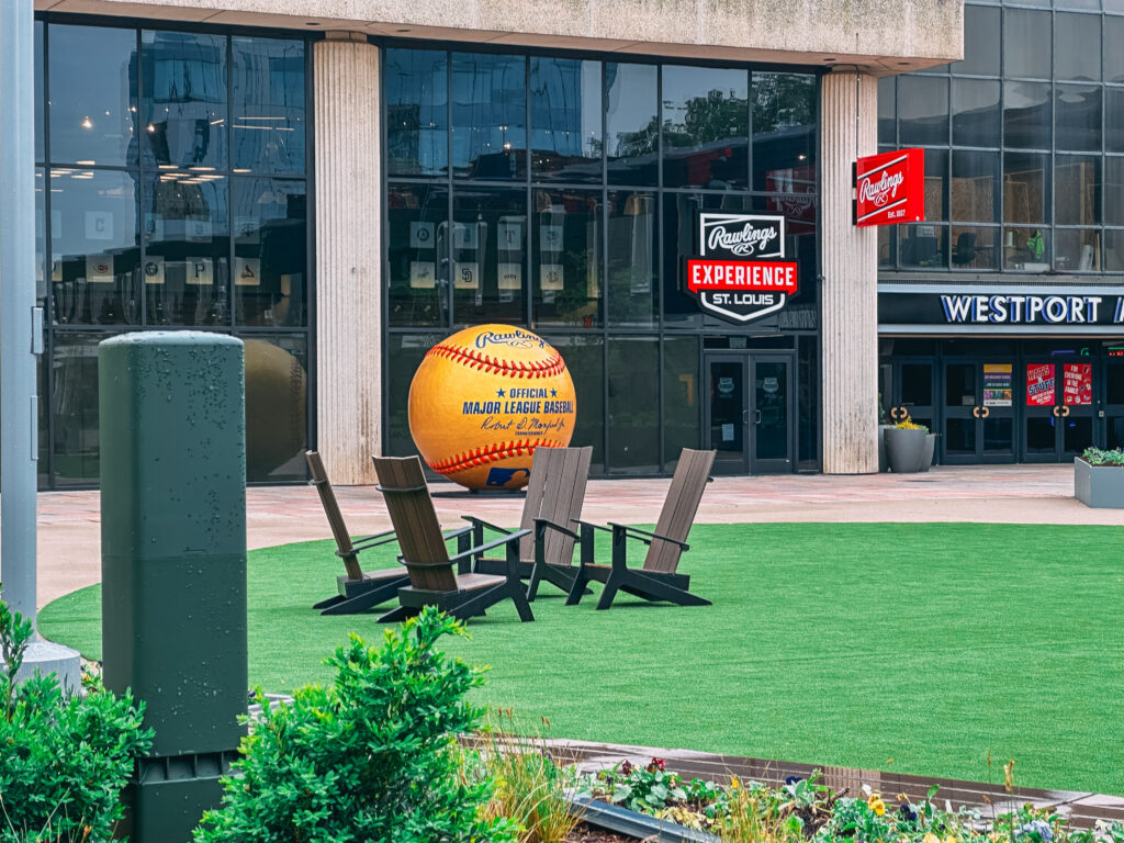 A plaza with Adirondack chairs on artificial grass sits in front of a building featuring a giant baseball and Rawlings signage for the Rawlings Experience St. Louis at Westport Plaza. Music for Business