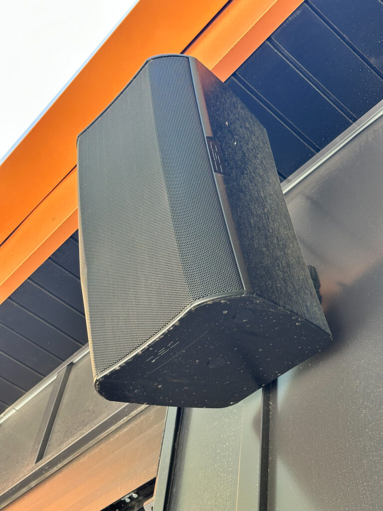 A close-up of a black outdoor speaker mounted on a wall under an orange roof overhang, with part of a dark ceiling and structural beam visible in the background. Music for Business