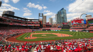Wide-angle view of a bustling baseball stadium filled with fans dressed in red. The field is lush and green, and the scoreboard is visible to the right. The city skyline and a famous arch are in the background under a blue sky with clouds.
