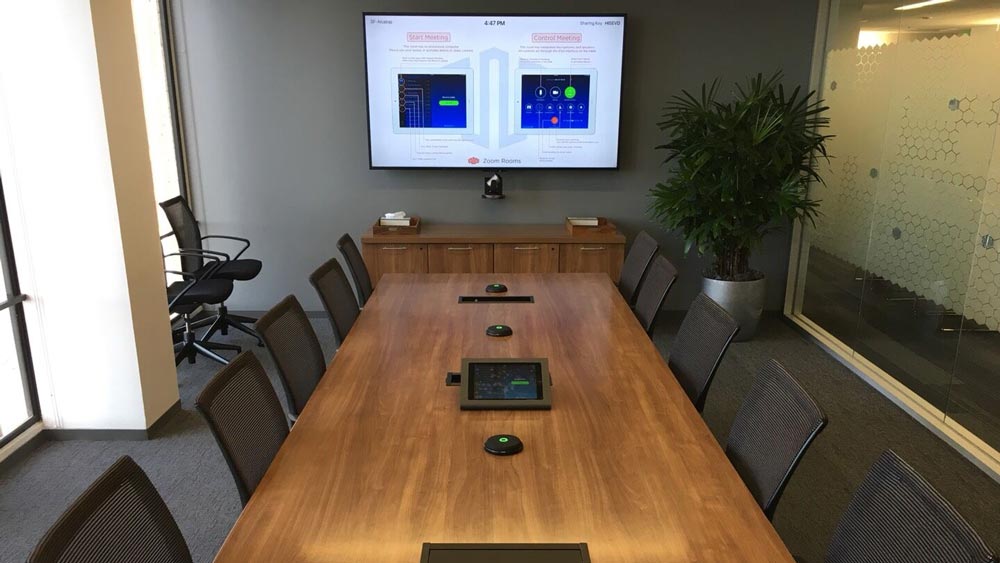 A modern conference room with a long wooden table, surrounded by black mesh chairs. A large screen at the front showcases graphs and data through advanced audio video solutions. Potted plants are in the corner, and a control tablet is on the table.