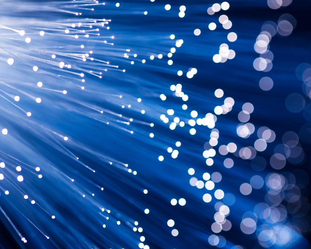 A close-up of glowing fiber optic cables intertwined with data voice cabling, showcasing bright blue and white lights. Beams of light spread out in different directions, creating a dynamic and futuristic effect.