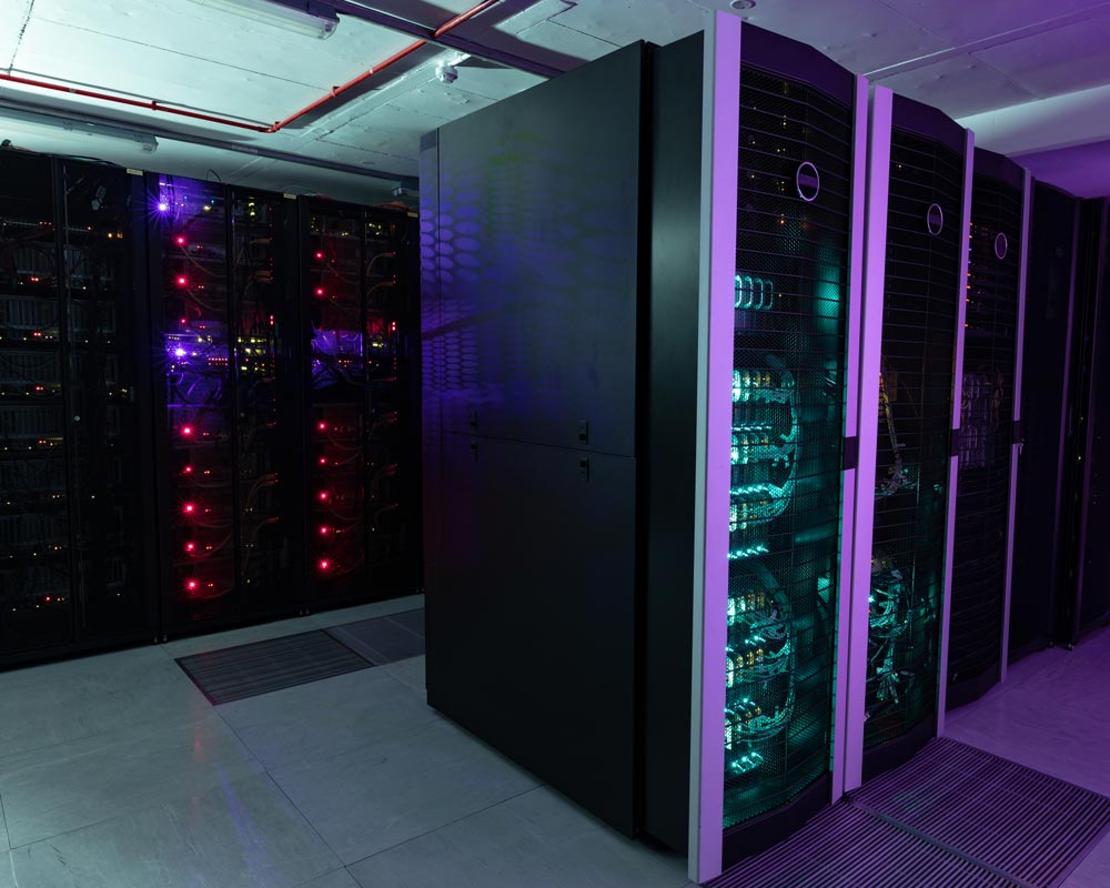 A dimly lit server room with rows of servers, their glowing red and blue indicator lights providing a futuristic glow, houses an intricate web of data cabling. Tall black cabinets line the walls in organized fashion, each concealing the pulse of the network within.