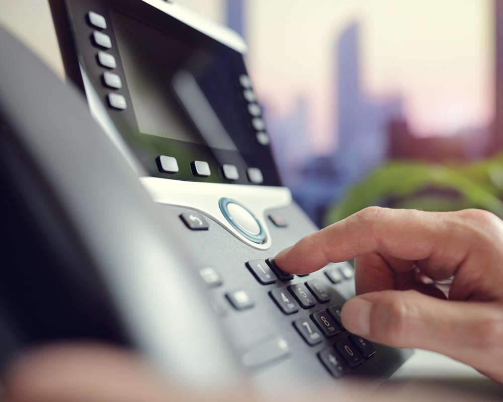 A person dialing a number on a modern office desk phone, seamlessly integrated with data voice cabling. The phone display and keypad are visible against a blurred cityscape in the background.