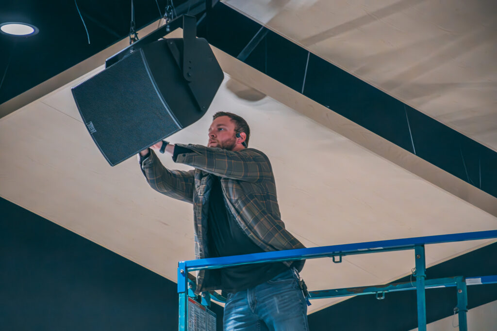 A person wearing a plaid shirt and jeans stands on a blue lift, adjusting a large black speaker suspended from the ceiling in an indoor setting.