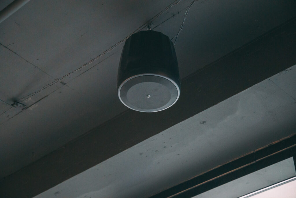 A black speaker is mounted on a gray ceiling with wires visible. The ceiling beams and shadows are apparent in the background.