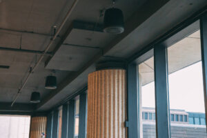 Interior view showing a row of large windows and ribbed concrete columns. Ceiling-mounted speakers are visible, and natural light illuminates the space. The ceiling has exposed beams and industrial-style fixtures.