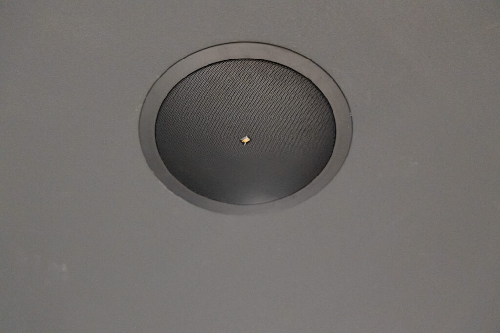 Ceiling-mounted circular speaker with a small central hole, set against a gray surface.