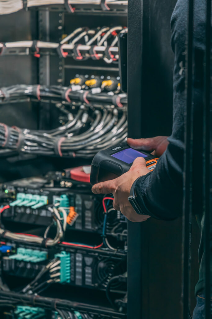 A person holding a device examines a server rack filled with cables and electronic equipment.