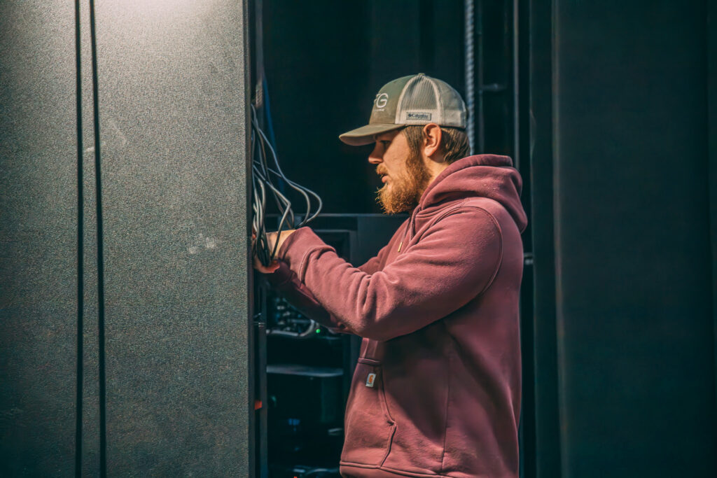 A man with a beard, wearing a gray cap and a maroon hoodie, adjusts cables on electronic equipment in a dark room.