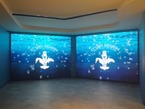 Two large digital screens display an underwater scene with bubbles and a cartoon otter holding a trident. The text reads "St. Louis Aquarium." The room has a tiled floor and curved ceiling.