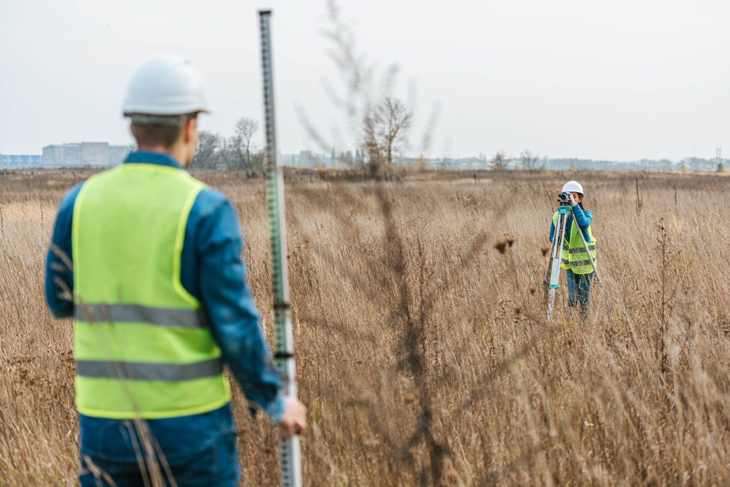 Two surveyors in reflective vests and helmets work in a grassy field. One holds a measuring rod, while the other looks through a survey instrument. The background shows a bare tree and distant buildings under a cloudy sky.