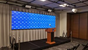 A press conference setup featuring a wooden podium with a microphone, placed on a raised platform. Behind the podium is a large blue backdrop with repeating logos. The room has black flooring and ropes cordoning off the area.