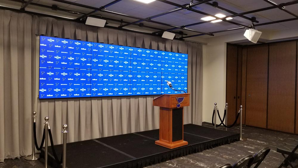 Empty podium on a small stage with a microphone, set in front of a large blue backdrop featuring a repeating logo. The room has a gray curtain, black carpet, overhead lights, and is flanked by retractable barriers with ropes. Chairs are arranged in front.