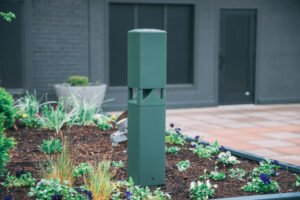 A tall, green outdoor speaker stands in a landscaped area filled with various plants and flowers. The background features a gray building with a black door.