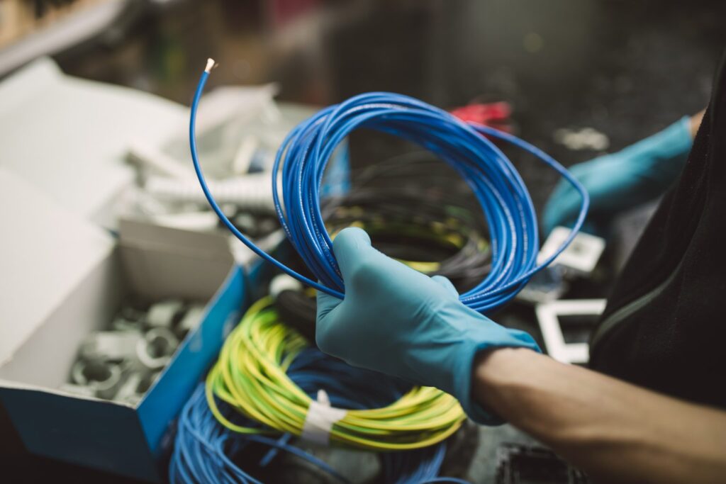 A person wearing blue gloves holds a coiled blue cable. Various cables and connectors are visible on a cluttered workspace, including green and yellow cables and a box containing hardware components.