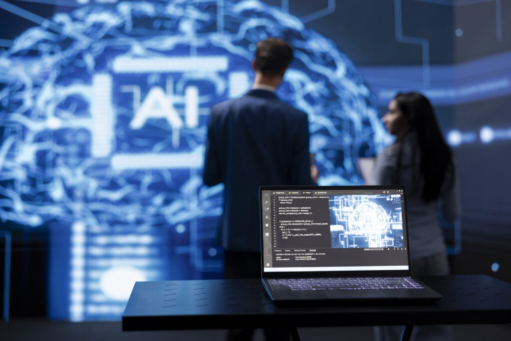 A laptop with code on the screen sits on a table in the foreground. In the background, two people stand in front of a large digital display showing a stylized brain and the letters "AI.