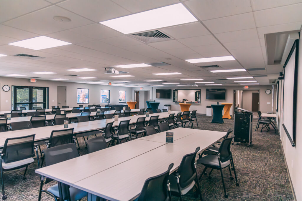 A spacious conference room with multiple rows of tables and chairs facing the front. The room has overhead lighting, large windows, and a few wall-mounted screens. The setup suggests readiness for a meeting or presentation. Voice Lift Technology