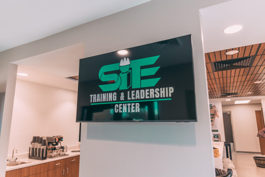 SITE-19 A television screen mounted on a gray wall displays the logo for "SITE Training & Leadership Center." The room includes a coffee station with machines and cups on a counter beneath recessed lighting. AV Solutions.