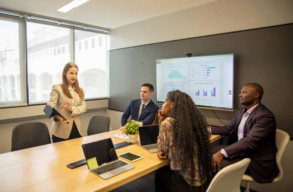 A woman stands and presents to three seated colleagues in a modern conference room. A wall-mounted screen displays graphs and charts. Laptops and a plant are on the table. Large windows allow natural light to illuminate the meeting space.