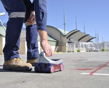 A person in work boots and blue pants places a small wheeled robot on a paved outdoor surface, with a modern building and clear sky in the background.