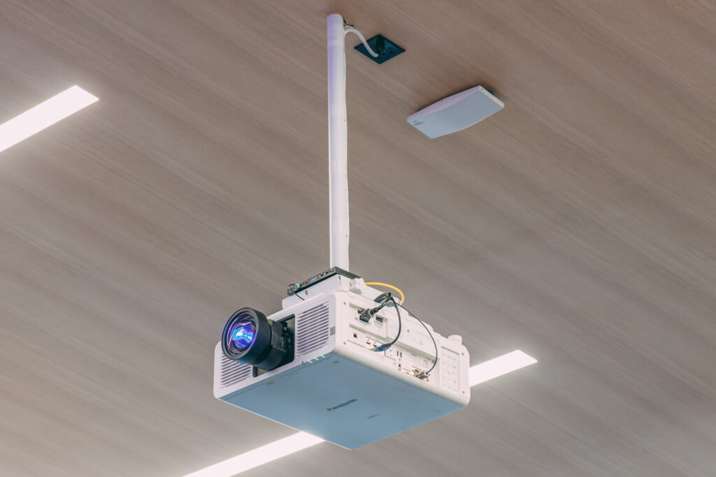 A ceiling-mounted projector, often used as municipalities use audio-video setups, with cables connected and attached to a white pole, is projecting blue light onto the wooden ceiling, which features a rectangular vent and a nearby small white device.