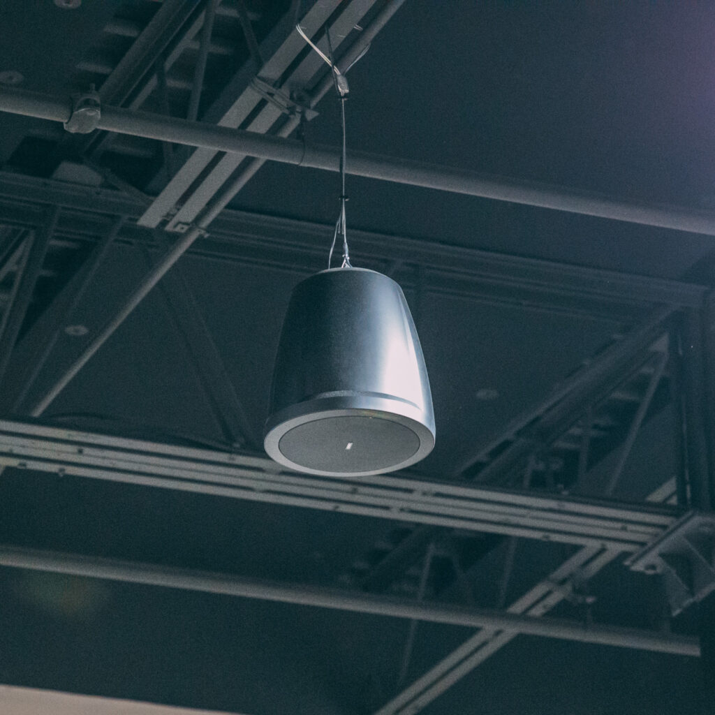 A black hanging speaker is suspended from an exposed industrial ceiling with visible metal beams and wires. Background music services
