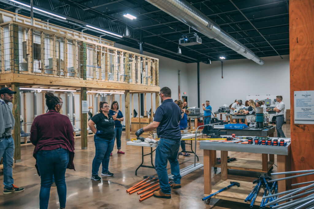 A group of people work and converse in a spacious workshop with tools, materials, and a large wooden structure. Some are gathered in small groups, while others are focused on individual tasks. The space is well-lit and organized.