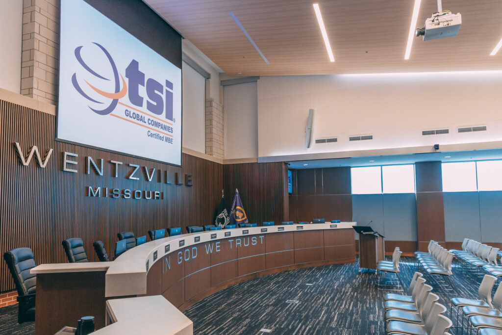 A modern city council chamber in Wentzville, Missouri, features empty seats, a curved desk, flags, and a large screen displaying the TSI Global Companies logo—demonstrating how municipalities use audio-video technology for meetings. "IN GOD WE TRUST" is visible on the desk.