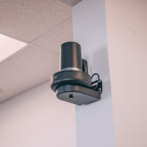 A black security camera is mounted high on a wall corner inside a building, positioned near a drop ceiling with white tiles. The camera is attached to a small black shelf with visible wires.