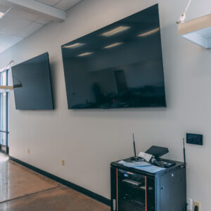 Two large flat-screen TVs are mounted on a light gray wall, and below them is a black equipment cart holding electronics, including a printer, router, and various cables. The room has a polished concrete floor.