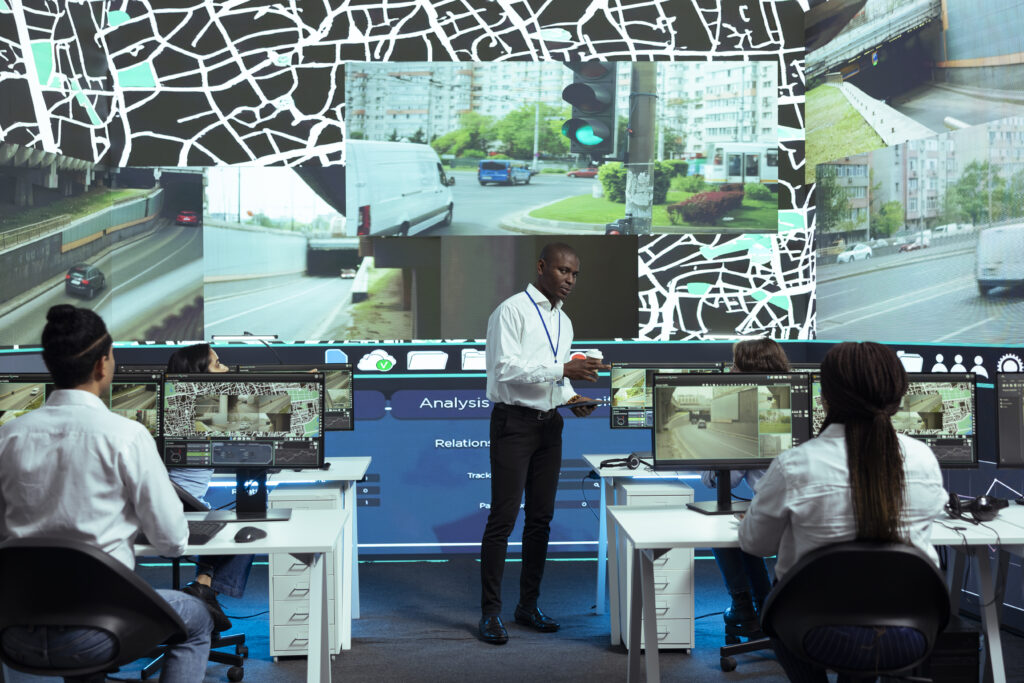 A group of people in a control room monitor traffic cameras and maps on large screens. One person stands and speaks while others sit at computers, all focused on analyzing vehicle and road surveillance data.