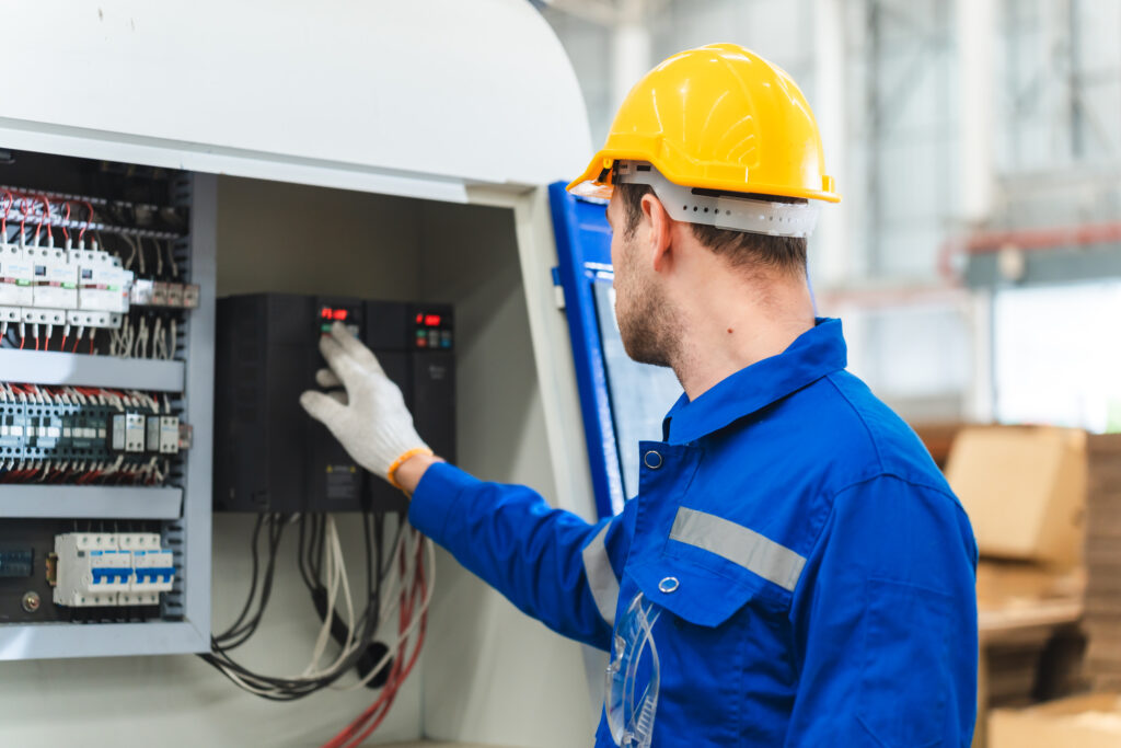 A worker in a blue uniform and yellow hard hat operates an industrial control panel, pressing buttons and adjusting settings inside an open electrical cabinet.