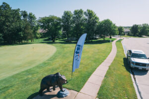 A bear statue and a promotional flag stand beside a golf course green near a sidewalk, with a white pickup truck parked on the grass and trees in the background under a clear blue sky.