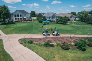 A golf course scene with two golf carts on a path, people gathered under blue tents on green grass, and large houses with patios and balconies in the background under a blue sky with scattered clouds.