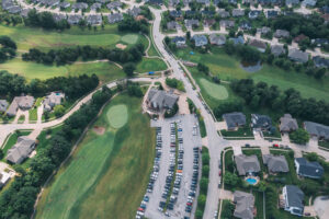 Aerial view of a suburban neighborhood with a golf course, a clubhouse, and a parking lot filled with cars; houses with lawns surround the area, and roads curve through the neighborhood.