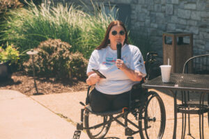 A woman in a wheelchair speaks into a microphone outdoors. She wears sunglasses, a light blue T-shirt, and holds a phone. A drink and table are beside her, with plants and a stone wall in the background.