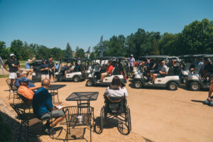 TSI Global Charity Golf Tournament, People gather outdoors near golf carts on a sunny day. Some are sitting at tables, including a person in a wheelchair, while others stand or prepare for golf. Trees and golf bags are visible in the background.