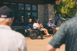 A small group of people, including a woman in a wheelchair, sit outside a stone building on a sunny day, while others stand nearby, some in hats, appearing to watch or listen to something off-camera.
