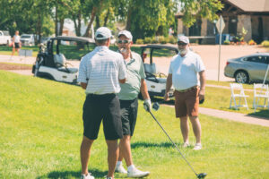 Three men on a golf course talk near golf carts; two are facing each other, one holding a club, while the third man stands nearby watching. Trees and a building are visible in the background.