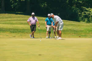 Four men stand on a sunny golf course green; one is putting while the other three watch. All wear casual golf attire and hats, with trees and grass visible in the background.