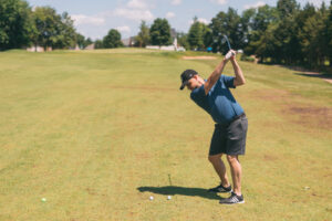 A man in a blue shirt and black shorts prepares to swing a golf club on a sunny golf course, with three golf balls on the grass in front of him and trees in the background.