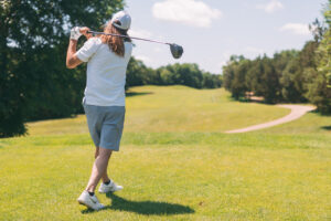 A person with long hair wearing a white cap, white shirt, and gray shorts swings a golf club on a sunny golf course, surrounded by green grass and trees under a blue sky.