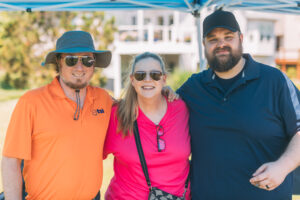 Three adults stand close together outdoors, smiling at the camera. The man on the left wears an orange shirt, sunglasses, and a sun hat. The woman in the middle wears a pink shirt and sunglasses. The man on the right wears a dark cap and a navy shirt.