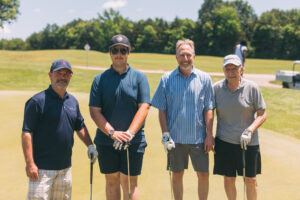 Four men stand together on a golf course, each holding golf clubs and smiling at the camera. It is a sunny day, and trees and grass are visible in the background.