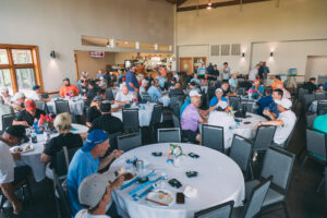 A large group of people sit at round tables in a bright, spacious room, socializing and eating food. The setting appears to be a casual indoor event or gathering, with plates and drinks on the tables.