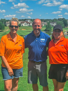 Three people stand smiling on a golf course. Two women wear orange shirts with a logo, and a man in the center wears a blue shirt. Trees, grass, and houses are visible in the background under a blue sky with clouds.
